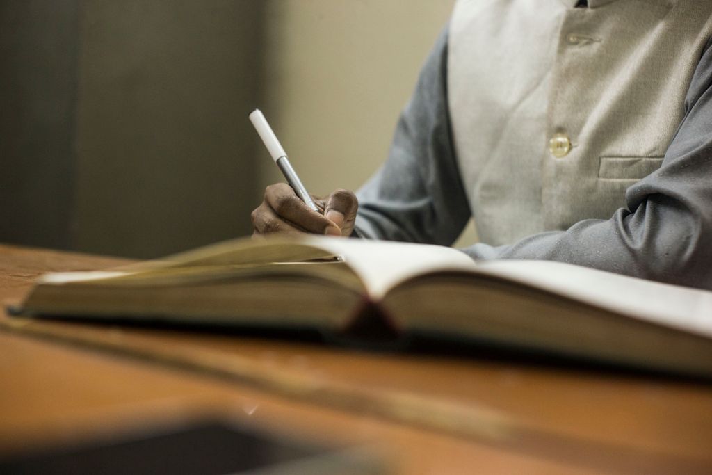 A person in a formal setting writes in a notebook at a wooden desk.